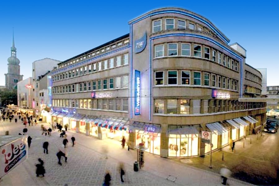 Eckgebäude mit Geschäften und beleuchteter Fassade, Passanten auf dem Platz